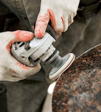 Stone fabrication worker in gloves and respirator grinding a granite slab, illustrating silica dust exposure risk in countertop manufacturing.